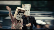 Szenenbild: Zwei Demonstrierende mit Tütenmasken vor einem verschwommenen Hintergrund